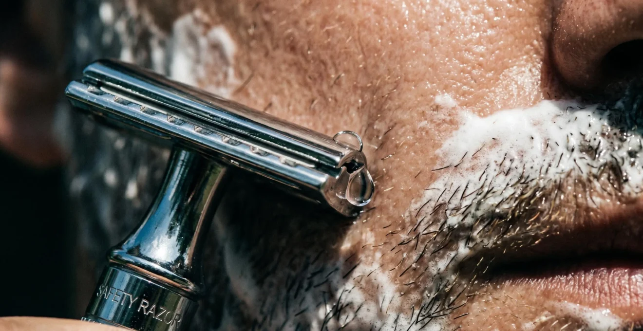 Gros plan sur une lame de rasoir formant le bon angle avec la peau lors du rasage, avec mousse et goutte d'eau.